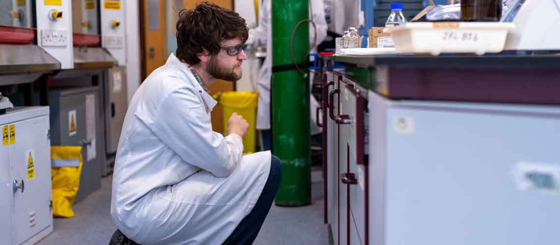 Researcher in a lab coat kneeling down to inspect bottles of liquids
