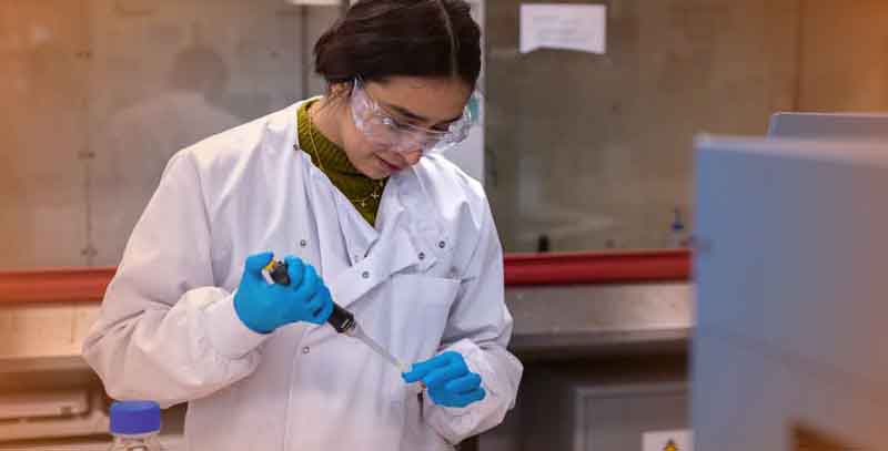 Female lab researcher injecting black liquid into a small tube while wearing protective glasses and gloves